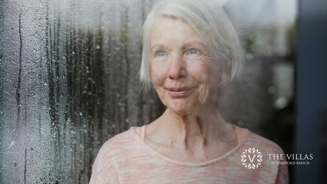 A senior woman smiling and watching the rain against a window.