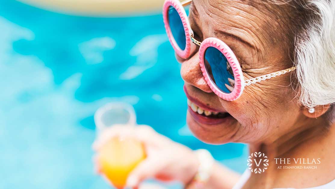 A senior woman wearing sunglasses and holding juice by the pool. Discover senior summer safety tips.